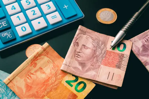 Moeda nacional (Real), uma caneta e uma calculadora em uma mesa escura - Repartição de receitas tributárias após a Reforma Tributária